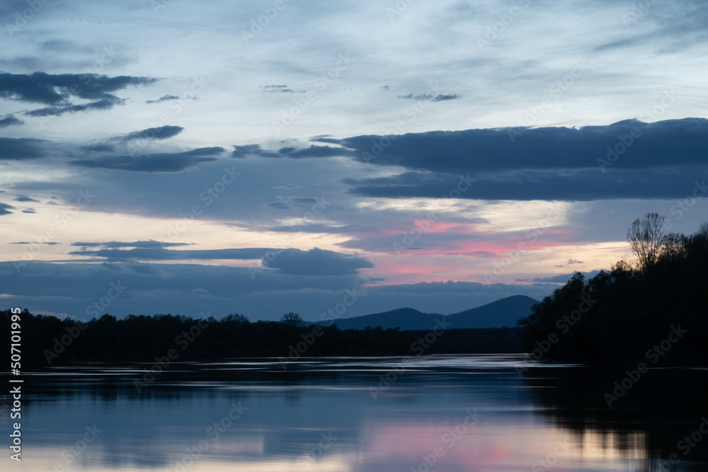 Landscape of river and mountain silhouette at dusk, Sava river with forested shore and Motajica mountain with clouds in sky during blue hour and purple glow in clouds
