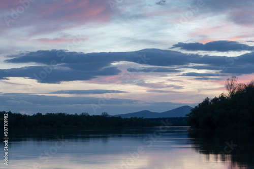 Landscape of river and mountain silhouette at dusk, Sava river with forested shore and Motajica mountain with clouds in sky during blue hour and purple glow in clouds