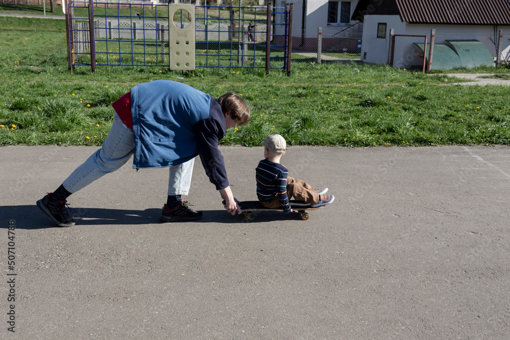 The older brother patiently and lovingly teaches the little boy his favorite hobby - to skate on a playground in the park in the fresh air. The older brother patiently and lovingly teaches the little boy his favorite hobby - to skate on a playground in the park in the fresh air.