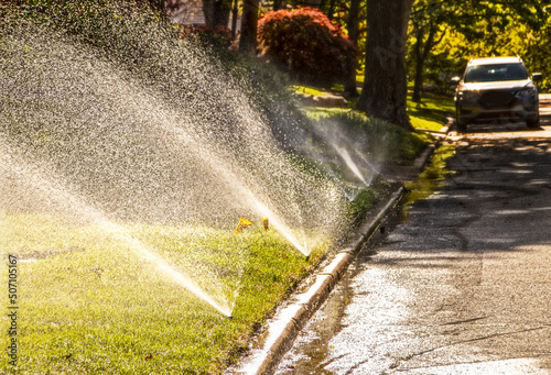 Sprinklers running in residential neighborhood with shady street with car parked in background - selective focus