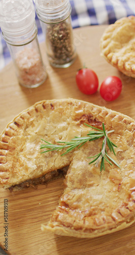 Vertical image of baked savoury pie missing slice, with rosemary, salt, pepper and tomatoes on table