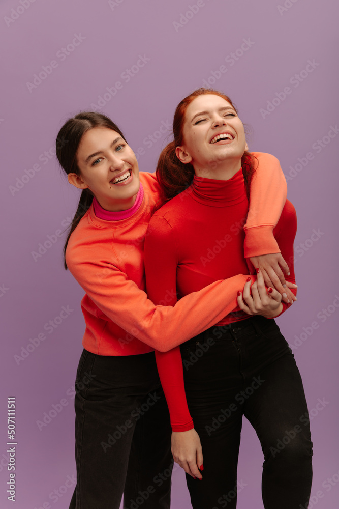 Obraz premium Pretty young caucasian girl hugging her sister standing in purple background. Teenagers in casual clothes laughing together. Lifestyle, different emotions, leisure concept. 