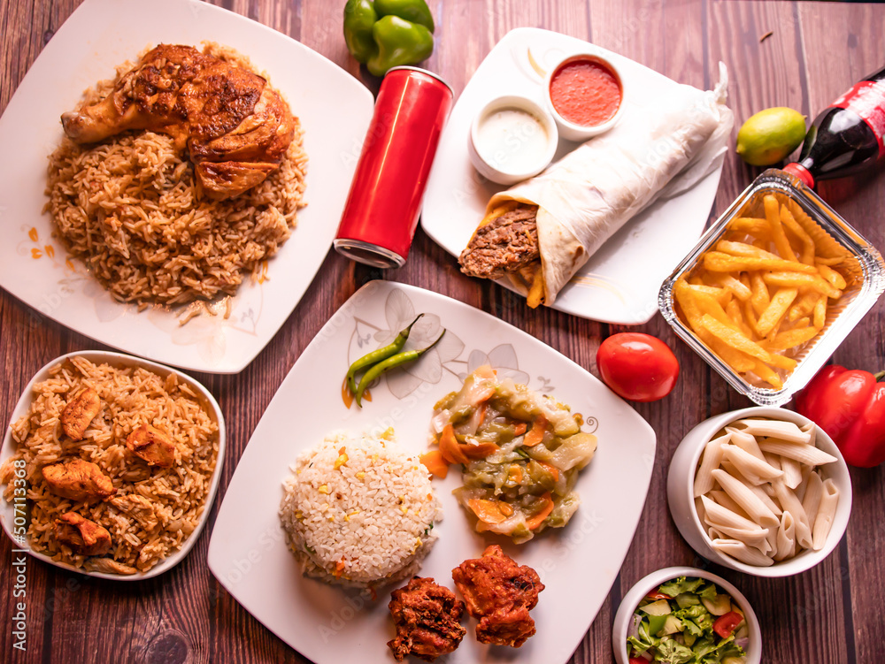 Assorted fast food table seekh kabab and tikka biryani, fried rice, fried chicken, seekh kabab shawarma, drink, french fries, raita and salad top view of wooden table