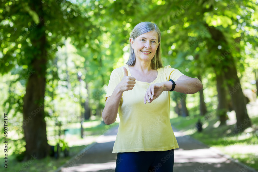 senior gray-haired retired woman in a summer park on a jog, looks at a smart watch and smiles, happy with the result senior gray-haired retired woman in a summer park on a jog, looks at a smart watch and smiles, happy with the result