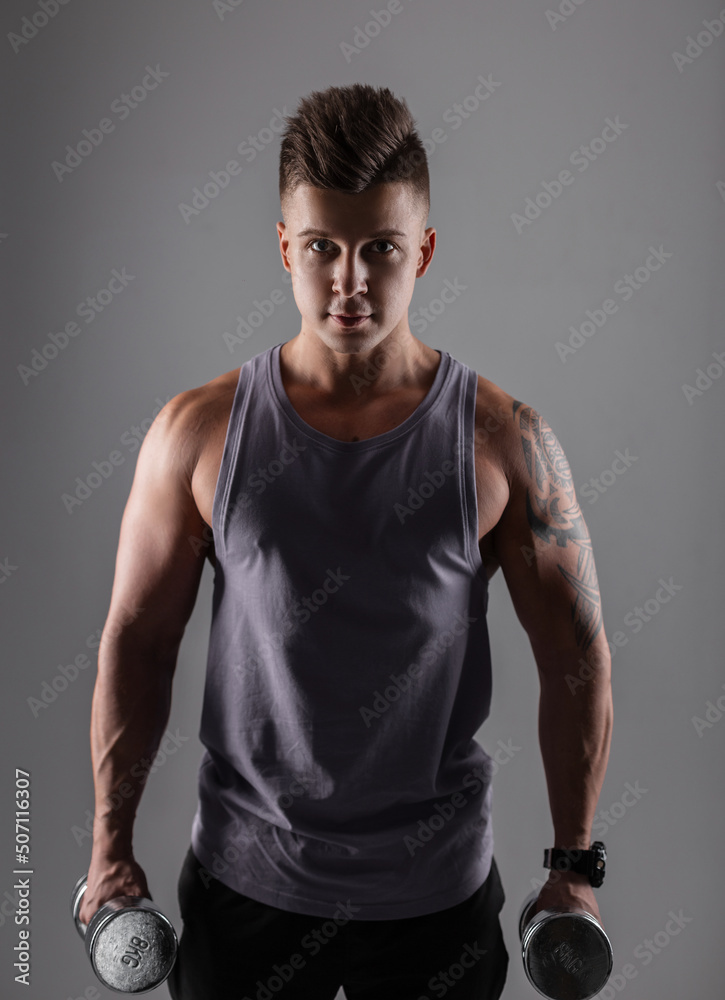 Fototapeta premium Handsome sporty young athletic man with shorty in sportswear with metal dumbbells on a dark background. Healthy lifestyle and workout