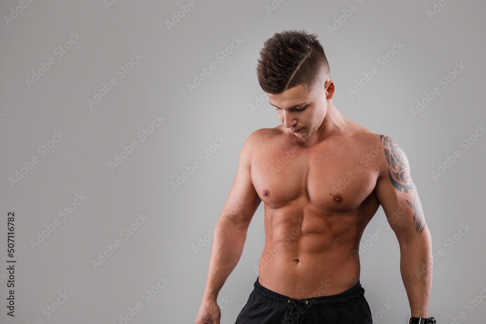 Male studio portrait of a handsome young athletic bodybuilder guy model with hair and a naked muscular sexy body with a tattoo on a gray background. Strong man model in studio