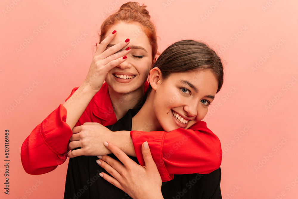 Close-up of young caucasian girl with her eyes closed hugging smiling friend from back against pink background. Brunette and redhead are wearing casual clothes. Concept of lifestyle emotions people. Close-up of young caucasian girl with her eyes closed hugging smiling friend from back against pink background. Brunette and redhead are wearing casual clothes. Concept of lifestyle emotions people.