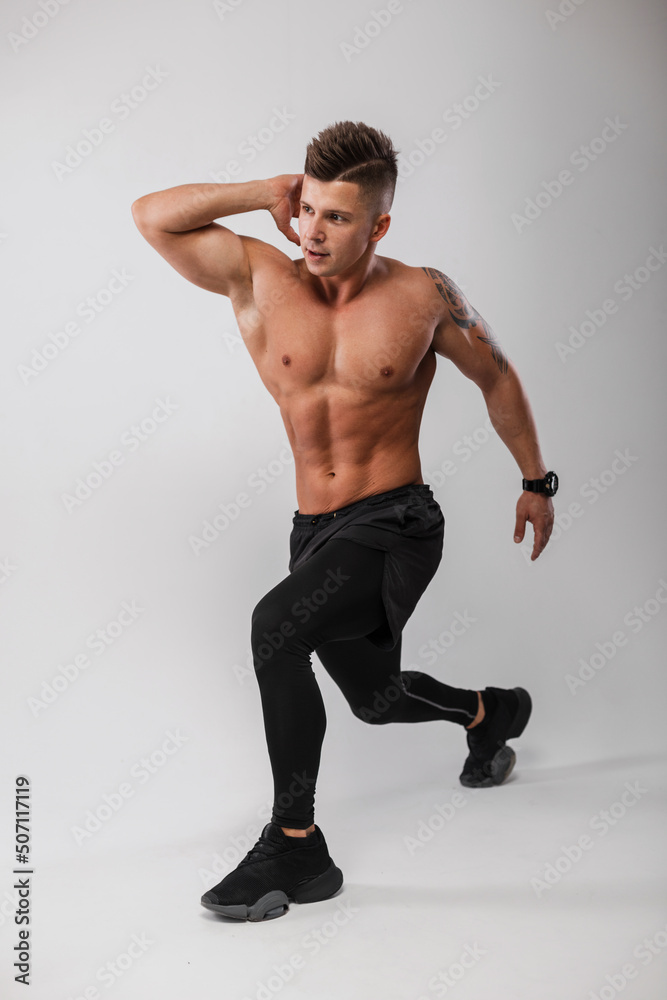 Handsome athletic man fitness model with a bare muscular torso in black sportswear with sneakers doing warm-up and exercise in the gray background Handsome athletic man fitness model with a bare muscular torso in black sportswear with sneakers doing warm-up and exercise in the gray background