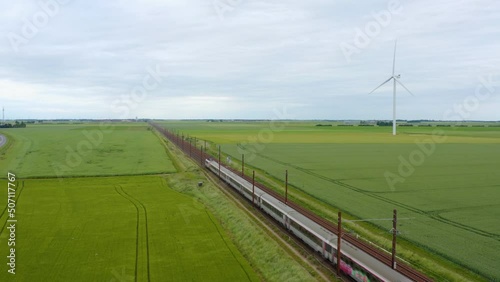 Aerial view of local train traveling through green fields under sunset sky and giant wind turbines. Train, railway in the meadow, windmills in the distance, sunset sky, Drone 4K. High-speed train. 