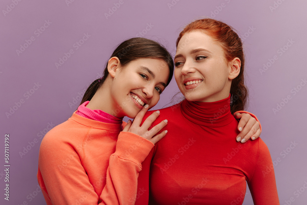 Beautiful young caucasian ladies with white smiles have nice time in purple background. Brunette looks at camera, redhead to side. Girlfriends are wearing bright sweaters. Lifestyle concept Beautiful young caucasian ladies with white smiles have nice time in purple background. Brunette looks at camera, redhead to side. Girlfriends are wearing bright sweaters. Lifestyle concept