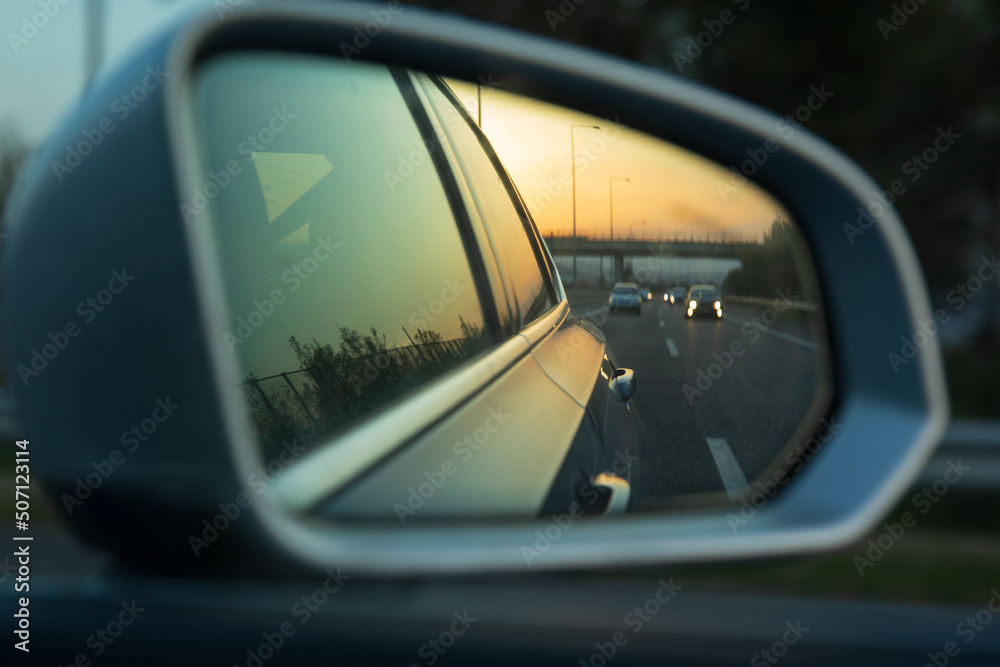 View of the outside car rearview mirror with reflections of the cars driving on the highway during sunset.