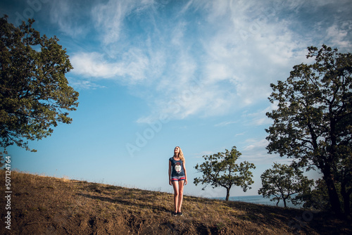 An attractive blonde girl in striped shorts and a blue t-shirt stands on the hills, around the trees, on a bright sunny day. There are many beautiful clouds in the sky.