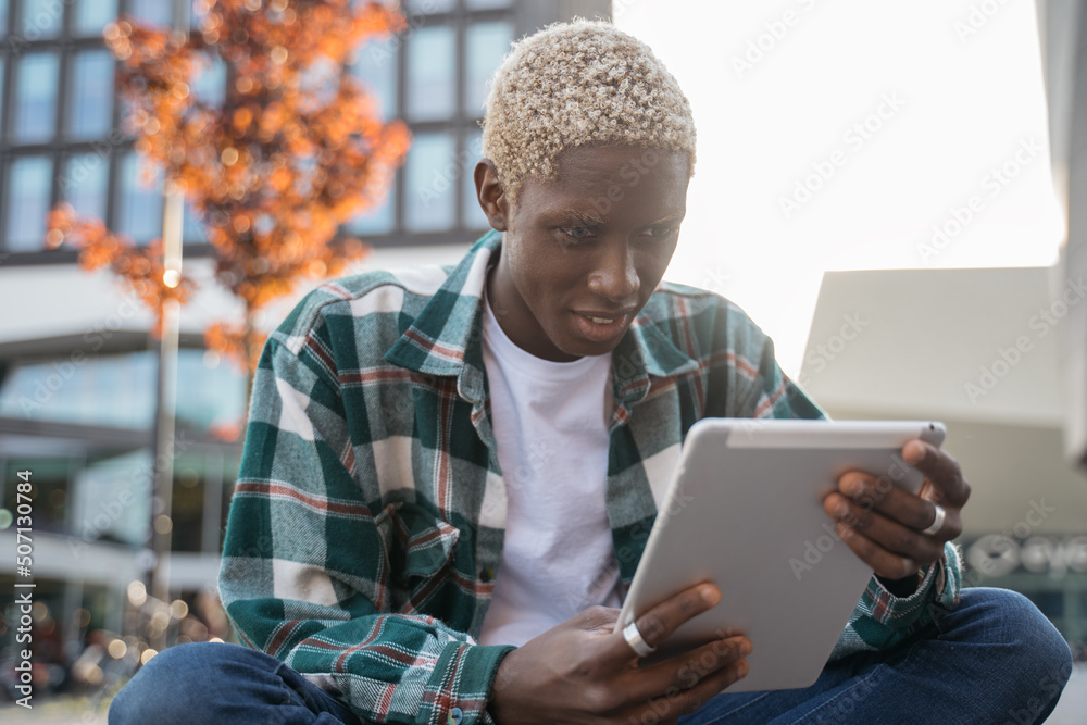 Obraz premium Young smiling African American student using digital tablet studying, learning language sitting in university campus, education concept. Hipster guy with stylish hair communication online outdoors 