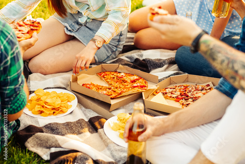 group of friends are enjoying pizza in the park. Men and women sit on the grass around a pizza box