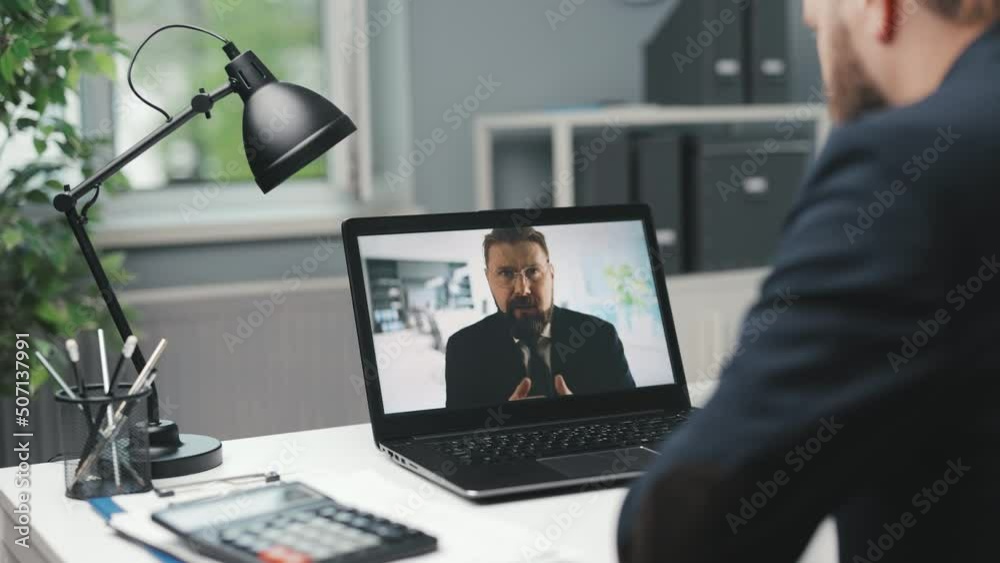 Back view of caucasian businessman having video call with his colleague while sitting at office desk. Two partners using modern laptop for online meeting. Back view of caucasian businessman having video call with his colleague while sitting at office desk. Two partners using modern laptop for online meeting.