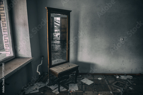 An old broken mirror stands in an abandoned building. Wooden mirror. Dirty and shabby walls. The depressing atmosphere of an abandoned building. Light from the window.