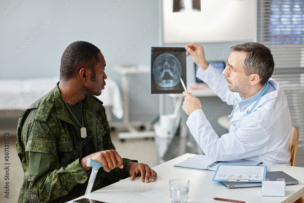 © Seventyfour - Professional medical worker showing skull x-ray picture to young adult military officer during appointment in hospital