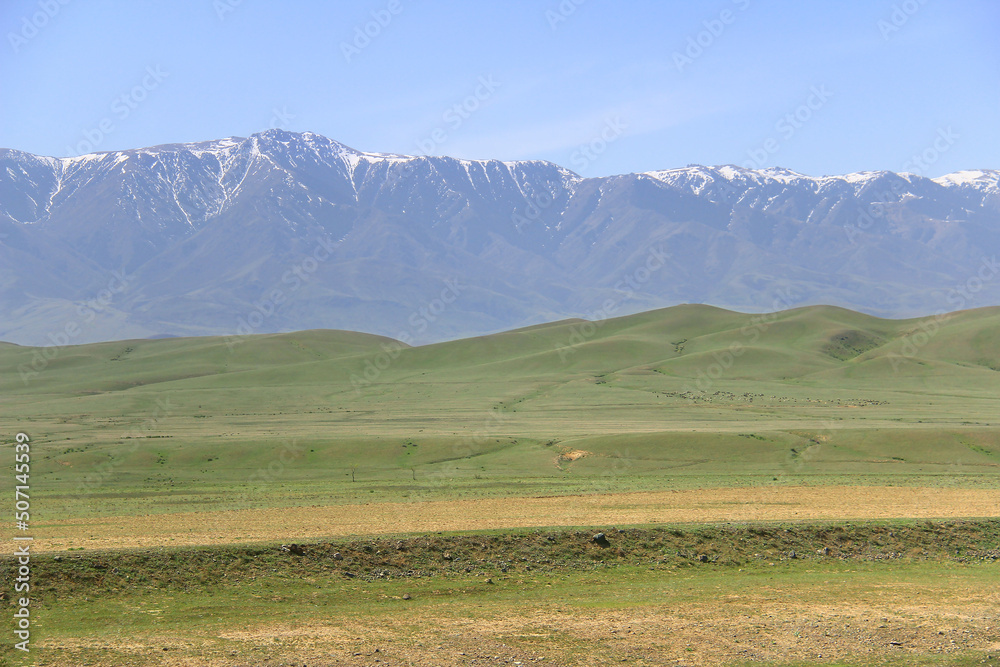 Big Alatau mountain range with snow-capped peaks, large green hills in the foreground, sky with clouds, spring, sunny