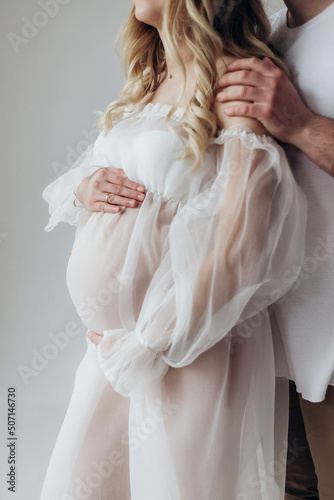 Husband and pregnant blonde wife in a white dress in the studio on a white background with a wreath on their heads hold their hands on their stomachs