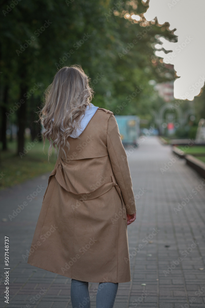 Young beautiful fair-haired girl in a sand-colored raincoat on a walk in the park in early autumn.