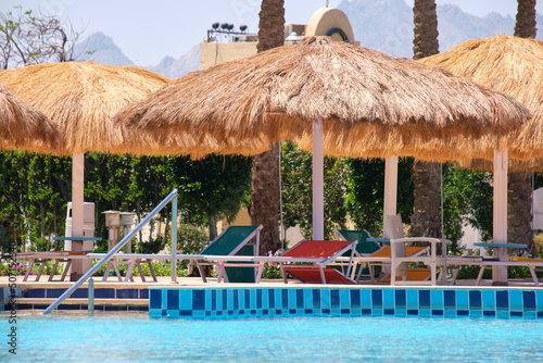 Empty deck chairs under straw shade umbrellas on swimming pool side in tropical resort. Summer vacations and getaway concept