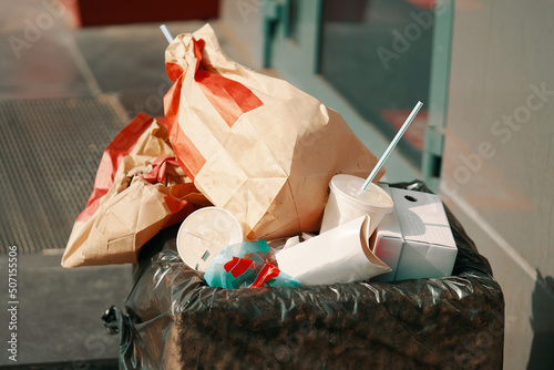 Bunch of paper packets and cups of coffee and other drinks in garbage. Dustbin next to fast food outlet. Urban landscape. Concrete trash can with black bag inside.