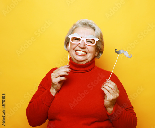 funny grandmother with fake mustache and glasses, laughs and prepares for party over yellow background