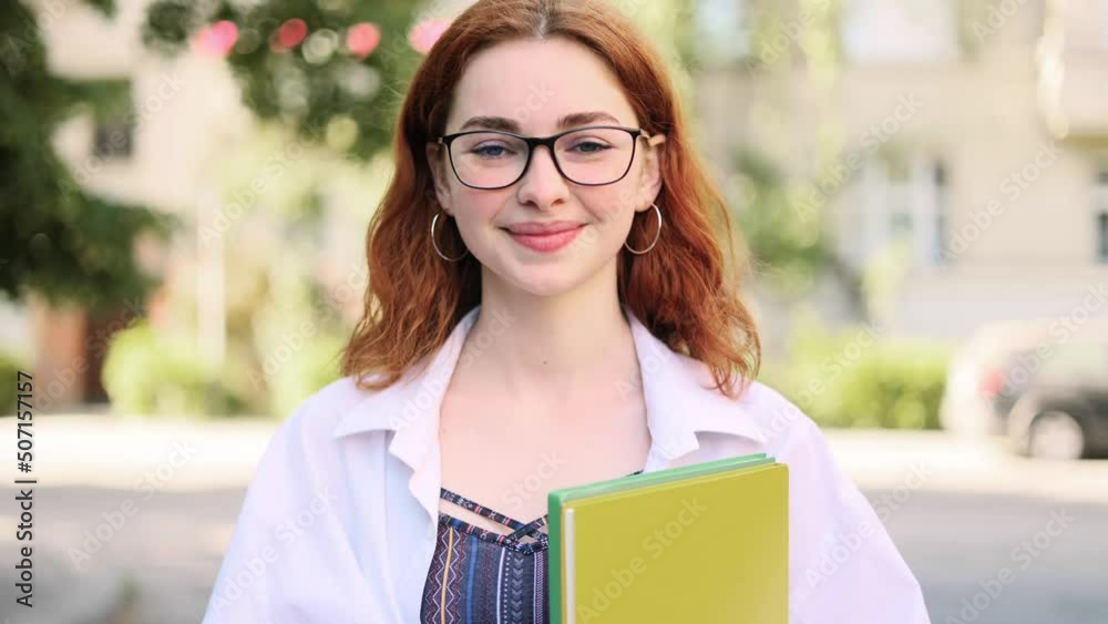 Student red head girl with eyeglasses standing with books near the park and looking at the camera. Portrait of attractive ginger girl. Student red head girl with eyeglasses standing with books near the park and looking at the camera. Portrait of attractive ginger girl.
