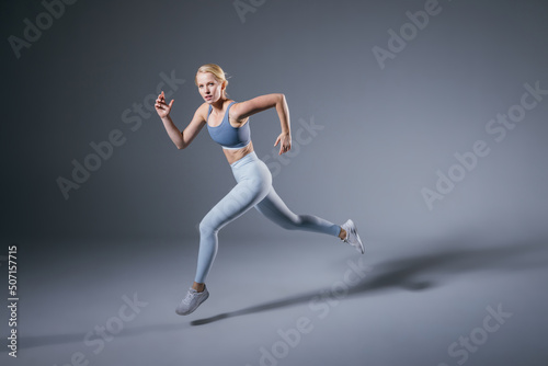Women running in studio. Female runner on blue gray background. Healthy blonde woman. Profile view of sport runner. Full length of fit girl