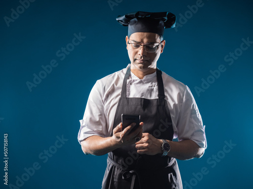 Man cook with phone. Guy in chef's uniform cook of restaurant on dark background. Career in restaurant concept. Indian restaurant owner. Chef in black cook hat. Cafe employee. Human confectioner photo