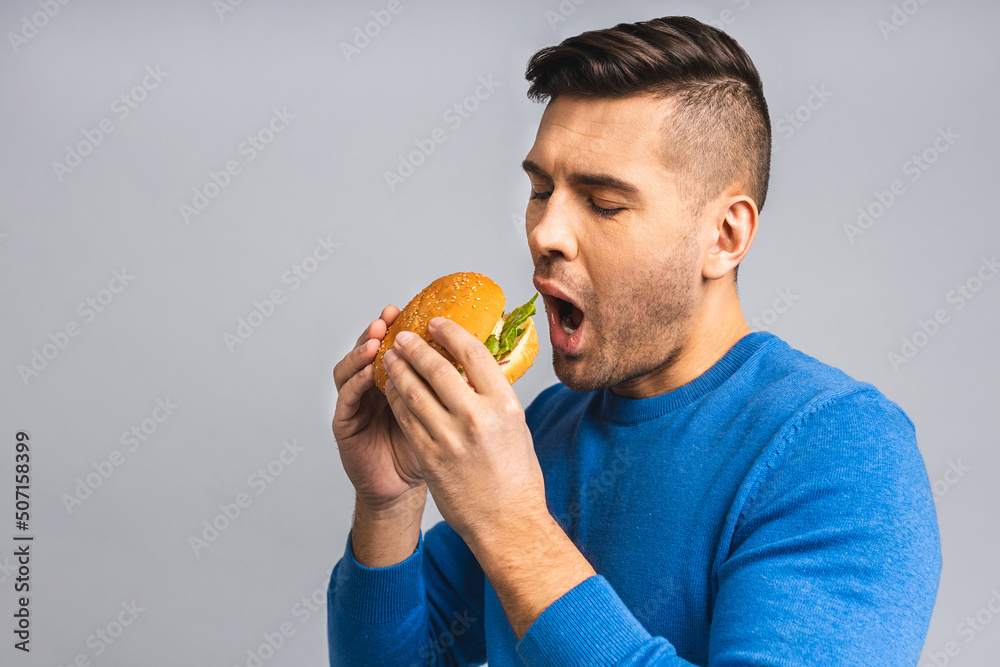 Young ukrainian man holding a piece of hamburger. Student eats fast food. Burger is not helpful food. Very hungry guy. Diet concept. Isolated over grey white background.