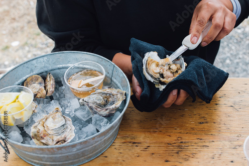 woman shucking oyster at table outdoors with ice-filled tub of oysters and condiments
