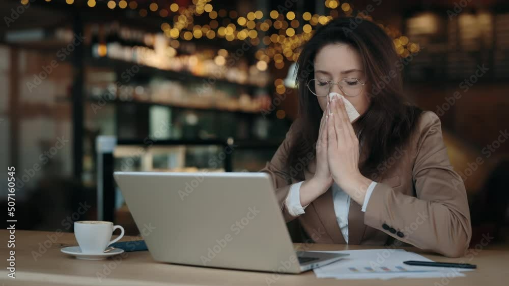 Sick business woman blowing nose in napkin while sitting at cafe and working on laptop. Caucasian lady in eyewear and formal jacket suffering from seasonal allergy. Sick business woman blowing nose in napkin while sitting at cafe and working on laptop. Caucasian lady in eyewear and formal jacket suffering from seasonal allergy.