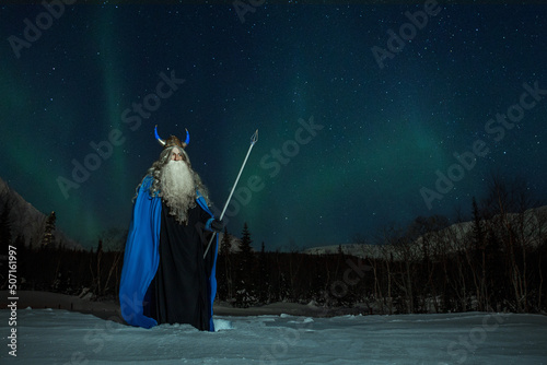 old man Odin with a staff stands in the snow at night against the backdrop of the northern lights