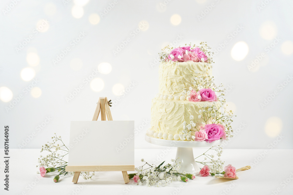 Wedding, Birthday 2 Tiered Cake for Cake Topper and Table Number Sign Easel Mockup. Styled with fresh pink roses and gypsophila flowers, against a white background with bokeh party fairy lights. Wedding, Birthday 2 Tiered Cake for Cake Topper and Table Number Sign Easel Mockup. Styled with fresh pink roses and gypsophila flowers, against a white background with bokeh party fairy lights.