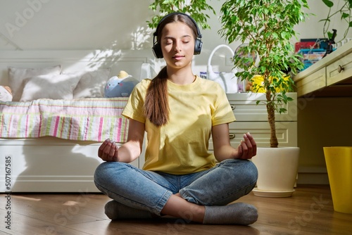 Teenage girl in headphones relaxing with closed eyes sitting in lotus position at home