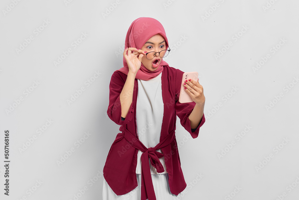 Surprised beautiful Asian woman in casual shirt looking at mobile phone, reading special offer, taking off glasses isolated over white background