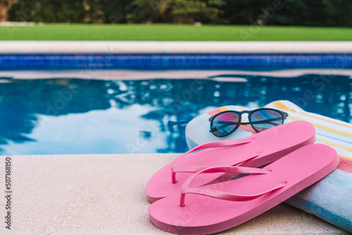 summery swimming objects in front of a swimming pool. beach towel, sunglasses, swimming flip-flops. lawn garden with swimming pool in the background.