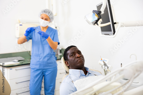 Pensive african-american man patient sitting on dental chair while Asian woman dentist standing in background and preparing tools for examination.