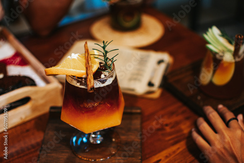 A glass of iced americano coffee and layer of orange juice decorated with rosemary leaf, cinnamon and sliced of orange on wooden coaster. Selective focus.

