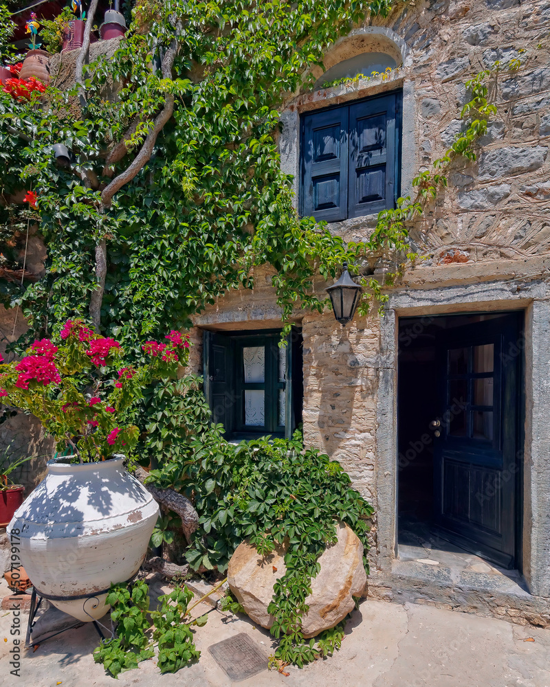A stone house with blue door and windows, a large jar and climbing flowers. Mesta village, Chios island, Greece. A stone house with blue door and windows, a large jar and climbing flowers. Mesta village, Chios island, Greece.