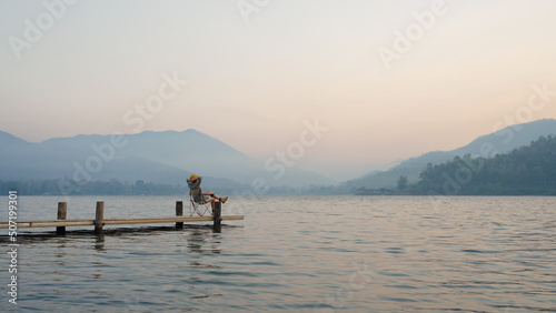 Asia people woman enjoy sit calm day dream on cozy chair at river pier hand behind head. Wide view of cloud dawn dusk sky solo budget staycation getaway at glamping take city life break stress relief.