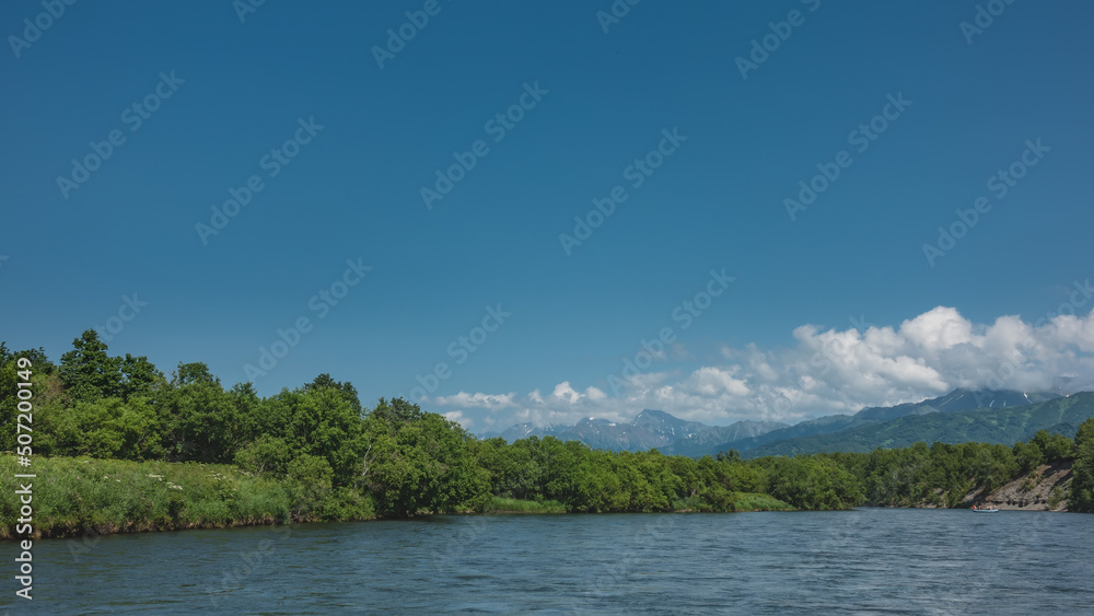 A calm blue river with  banks overgrown with lush vegetation. An inflatable rafting boat with people is visible in the distance. A picturesque mountain range against the sky and clouds. Kamchatka. 