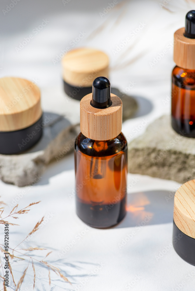 Amber glass dropper with bamboo lid and black jars. Bottle on grey concrete podium for product presentation. Skincare cosmetic with wildflowers. Beauty concept Amber glass dropper with bamboo lid and black jars. Bottle on grey concrete podium for product presentation. Skincare cosmetic with wildflowers. Beauty concept