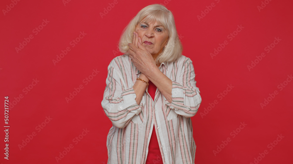 Senior elderly woman touching sore cheek suffering from toothache cavities or gingivitis waiting for dentist appointment gums disease. Mature grandmother indoors studio shot isolated on red background