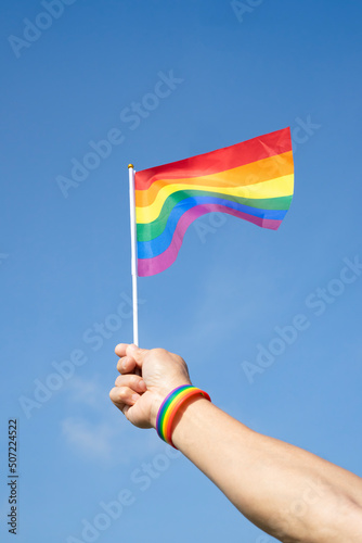 man s hand wears LGBT rainbow wristband is waving LGBT rainbow flag on background blue sky selective focus   concept for LGBTQ  equality movement community