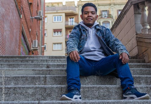 full length body image of a short Mexican man sitting with his hands leaning on his knees in the steps of a neighborhood