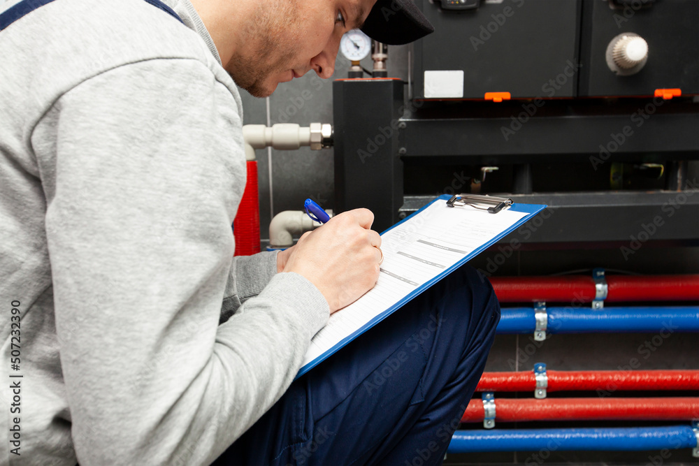 Technician servicing holding clipboard and inspecting heating system in boiler room