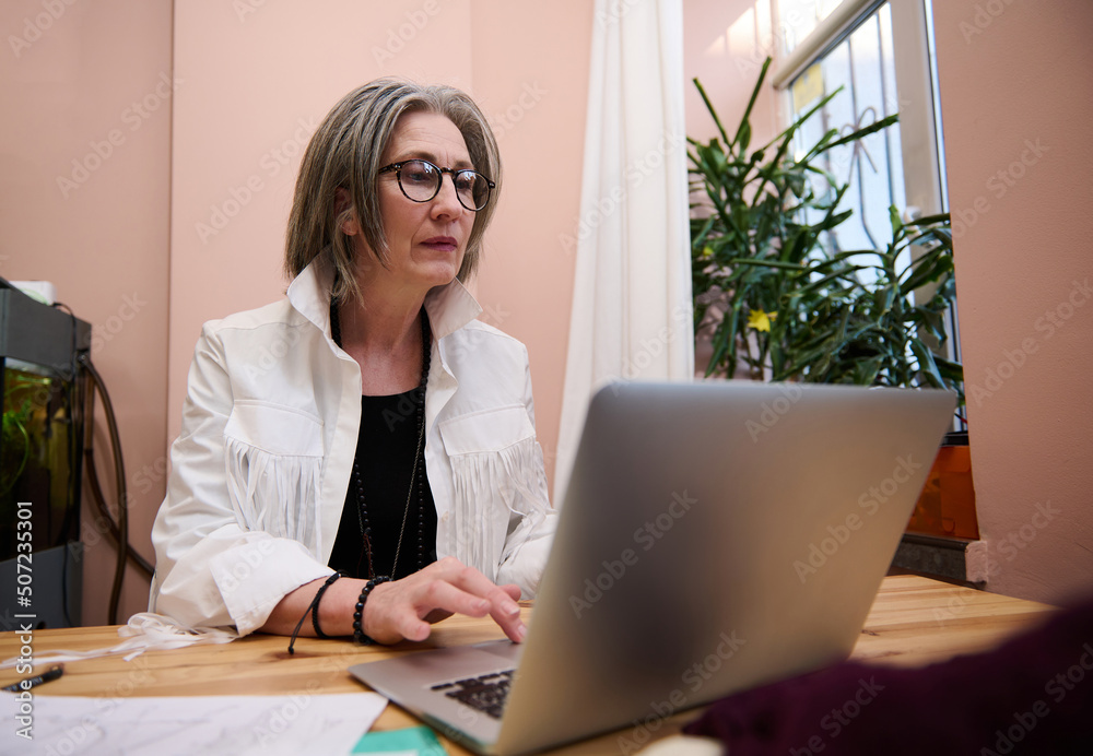 Elderly European woman fashion designer working in clothing design and tailoring studio and using laptop while designing new garment Elderly European woman fashion designer working in clothing design and tailoring studio and using laptop while designing new garment