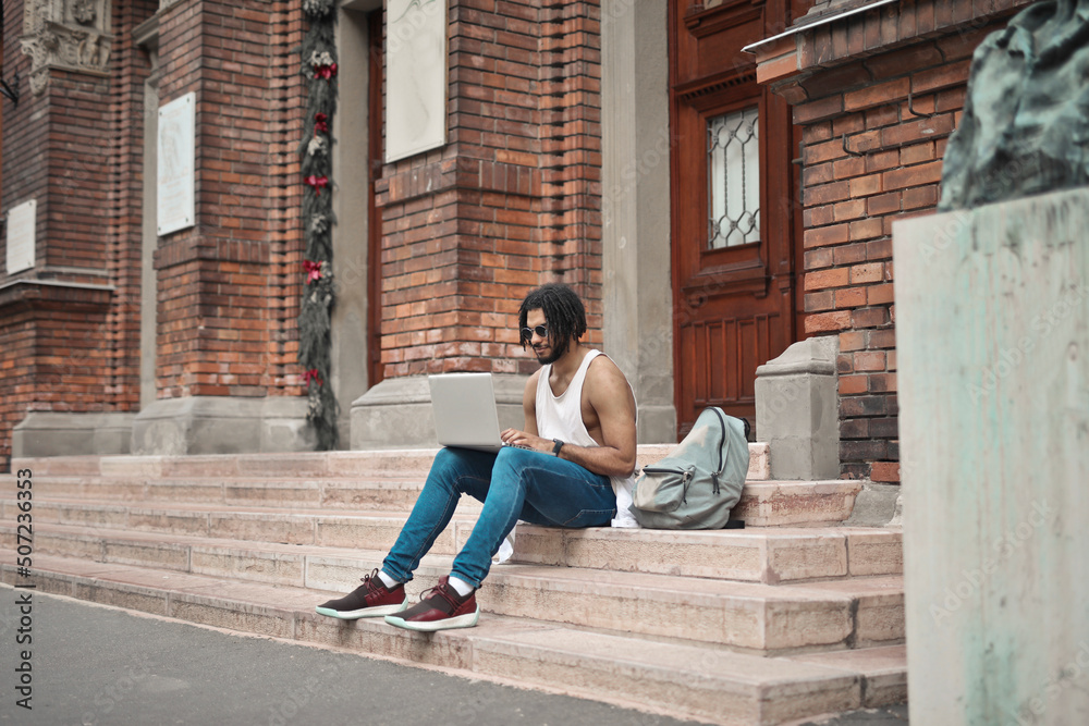 young man sitting on the steps of a school entrance uses a computer young man sitting on the steps of a school entrance uses a computer